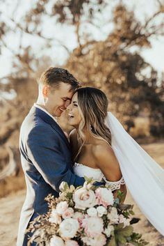 a bride and groom embracing each other in front of some trees with their wedding bouquet