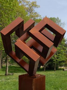 an abstract sculpture in the middle of a grassy area with trees and bushes behind it