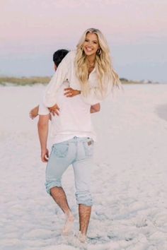 a man carrying a woman on his back at the beach while she walks in the sand