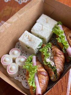 an open box filled with sandwiches and other food on top of a wooden table next to rolls