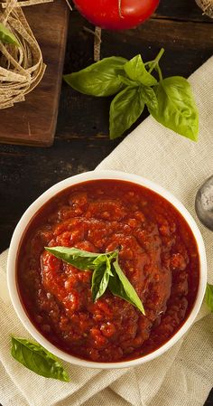 a bowl of tomato sauce with basil leaves on the side and tomatoes in the background