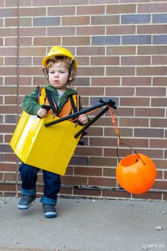 a little boy dressed up as a construction worker holding a bucket and shovel while standing in front of a brick wall