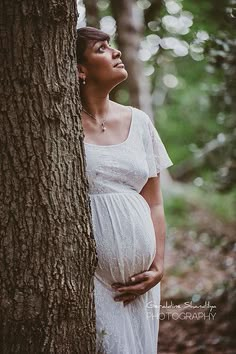 a pregnant woman leaning against a tree in the woods