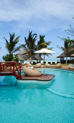 an outdoor pool with lounge chairs and umbrellas next to the ocean in front of palm trees