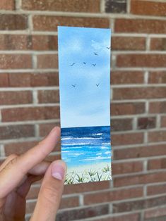 a person holding up a piece of paper with water and birds flying over the ocean