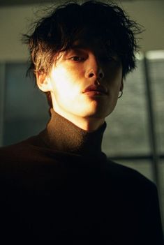 a young man with dark hair standing in front of a window wearing a black turtle neck sweater