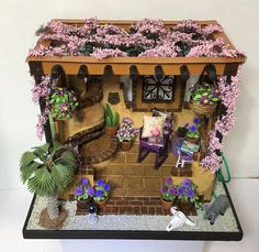 a miniature house with flowers and plants on the front porch, sitting next to a brick wall