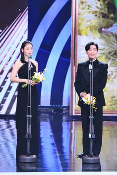 two people standing on stage with microphones and flowers in front of them, one holding a bouquet