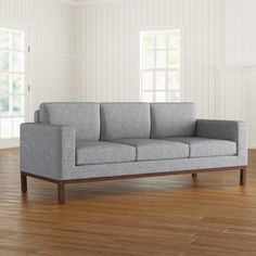 a gray couch sitting on top of a wooden floor next to two large windows in a white room