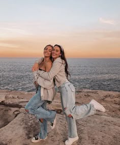 two women hugging each other while standing on rocks near the ocean at sunset or sunrise