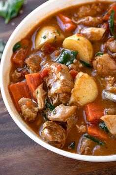 a white bowl filled with meat and vegetables on top of a wooden table next to a spoon