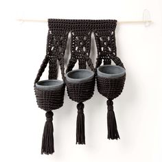 three black crocheted pots hanging on a wall with tassels attached to them