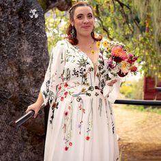 a woman in a white dress holding flowers and standing next to a tree with her hands on her hips