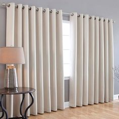 a living room with white curtains and a table in front of the window that has a lamp on it