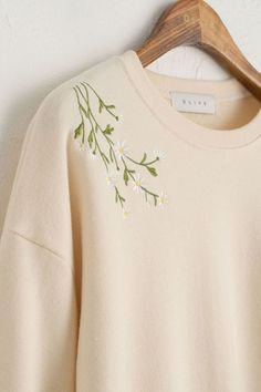 a white sweater with flowers embroidered on the front and back, hanging from a wooden hanger