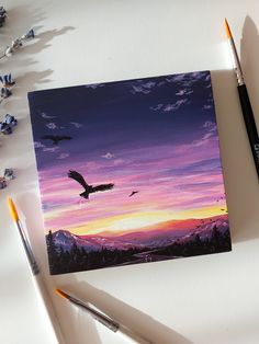 a painting of a bird flying in the sky over mountains at sunset with paintbrushes next to it