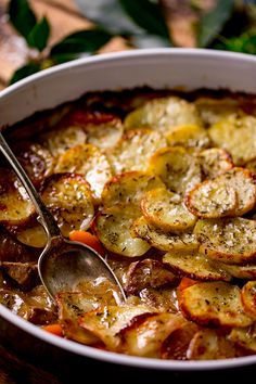 a white bowl filled with potatoes and meat on top of a wooden table next to a spoon