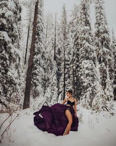 a woman in a purple dress sitting in the snow with trees behind her and she is wearing black