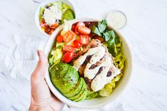 a person holding a bowl filled with salad and dressing on top of a white table