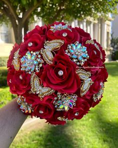 a bridal bouquet made with red roses and brooches is held in front of a house