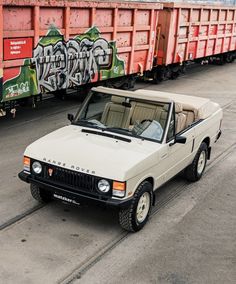 an old white car parked in front of some train cars with graffiti on the side