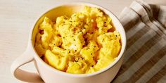 a white cup filled with scrambled eggs on top of a wooden table next to a striped napkin