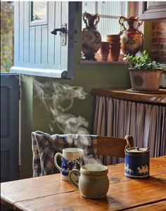 a wooden table topped with two mugs filled with steaming liquid next to a window