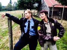 two people standing next to each other in front of a fence with a barn behind them