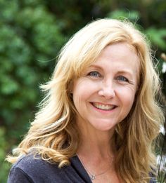 a woman with blonde hair and blue eyes smiles at the camera while standing in front of some trees