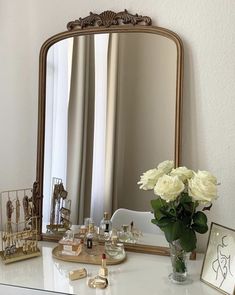 a white dresser topped with a vase filled with flowers next to a mirror and perfume bottles