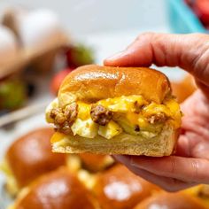 a hand holding up a sandwich with eggs and sausage on it in front of other muffins