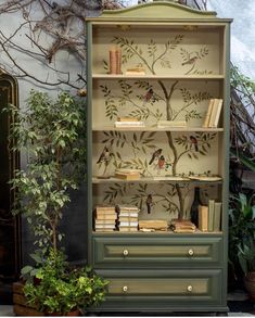 a green bookcase with birds and branches painted on the side, sitting next to a potted plant