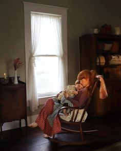 a woman sitting in a rocking chair holding a teddy bear and looking out the window