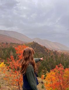 a woman standing on top of a hill pointing at the trees with orange and yellow leaves