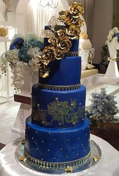 a three tiered blue and gold wedding cake on a white table with flowers in the background