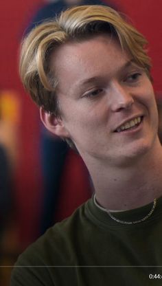 a young man with blonde hair smiles at the camera while wearing a green t - shirt