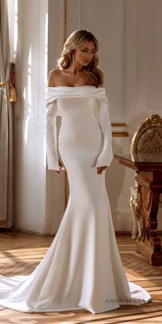 a woman in a white wedding dress standing next to a chair and window with her hands on her hips