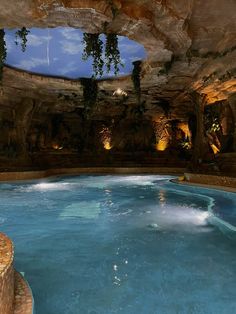 an indoor swimming pool surrounded by stone walls and plants on the ceiling is lit up at night