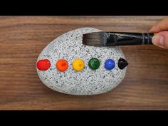 a hand holding a paintbrush over a painted rock with seven colored dots on it