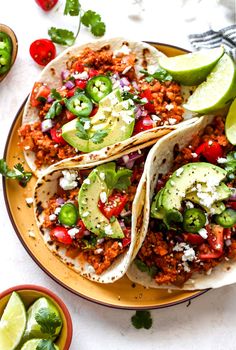 three tacos on a plate with limes and tomatoes