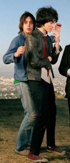 two young men standing next to each other on top of a hill with the city in the background