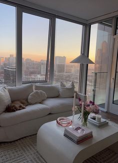 a living room filled with white furniture and tall buildings in the background at sunset or dawn