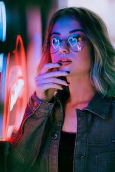 a woman wearing glasses standing in front of neon lights with her hand on her chin