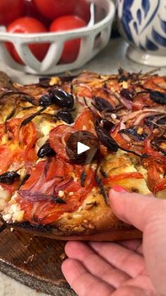 a person is holding up a pizza with tomatoes in the bowl behind it on a table