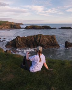 a woman sitting on top of a lush green hillside next to the ocean and rocks