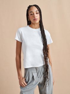 a young woman with long hair standing in front of a beige wall wearing grey pants and a white t - shirt