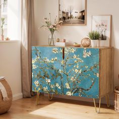 a blue cabinet with white flowers painted on the side and gold legs, in front of a window