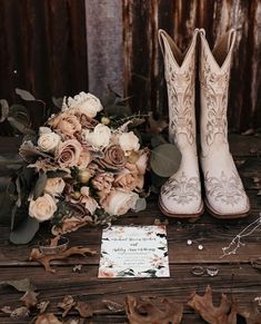 a bouquet of flowers and cowboy boots are sitting next to a wedding card on a wooden table