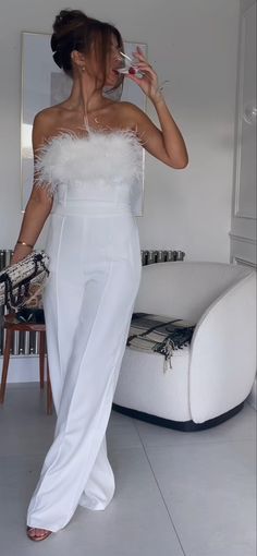 a woman in a white dress drinking from a wine glass while standing next to a couch