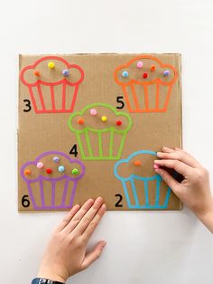 two hands on a piece of cardboard with colored cupcakes and numbers drawn on it